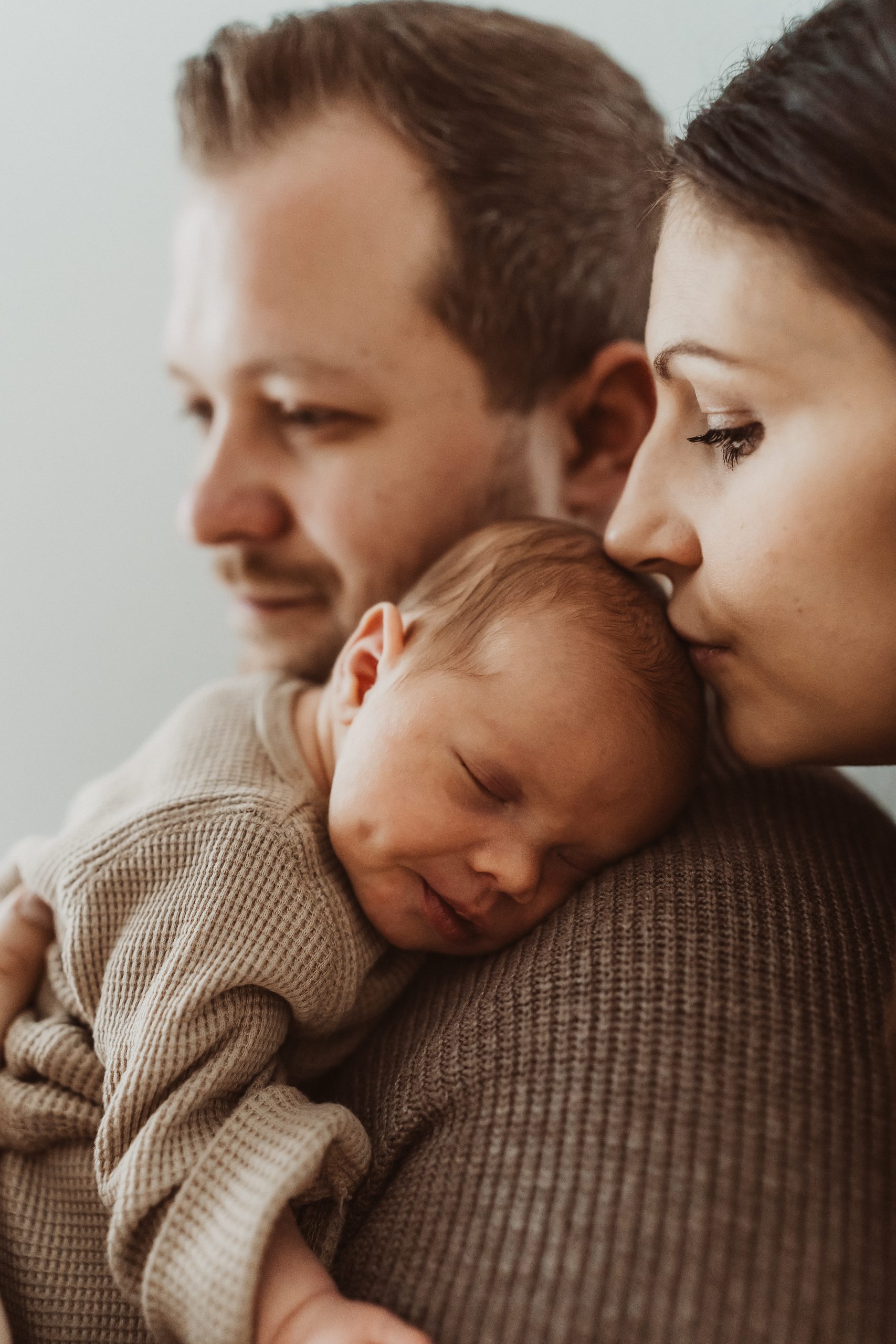 Der Vater hält das Baby auf der Brust. Während die Mutter dem Baby über der Schulter des Vaters einen Kuss auf die Stirn gibt